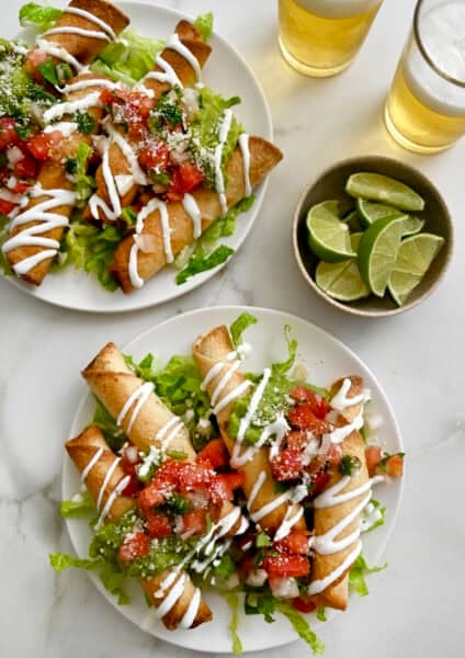 Two plates containing crispy chicken taquitos atop a bed of shredded lettuce and topped with pico de gallo and a drizzle of sour cream.