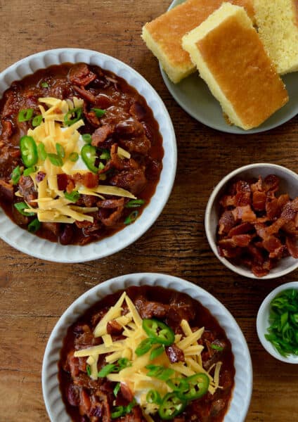 Top-down view of bowls of Chili con Carne topped with, sour cream, shredded cheese and jalapeños next to a small bowl containing pieces of bacon and a plate with cornbread