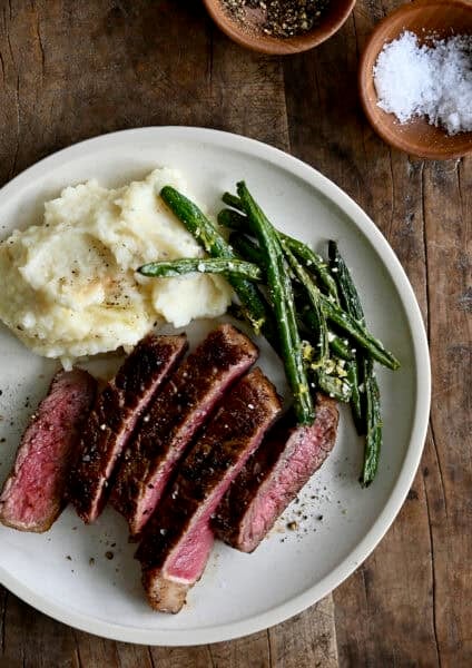 Sliced pan-seared steak served on a plate with blistered green beans and creamy mashed potatoes, showing a juicy pink center and golden crust.