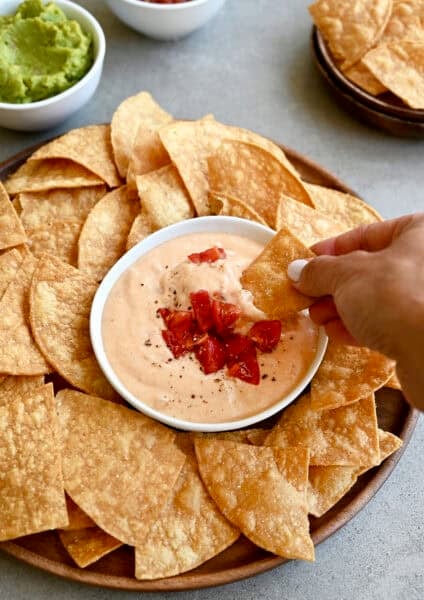 High-protein cottage cheese queso dip in a bowl topped with roasted tomatoes and black pepper on a plate surrounded by tortilla chips.