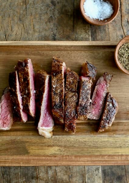 Sliced pan-seared steak on a wooden cutting board with small bowls containing salt and pepper nearby.