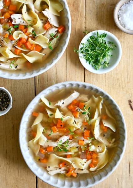 Two bowls containing chicken noodle soup next to a small bowl containing micro greens.