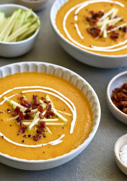 Two bowls containing butternut squash apple soup garnished with apple matchsticks, crispy pancetta and a drizzle of sour cream.
