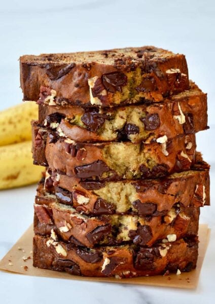 Slices of banana bread stacked on top of each other with bananas in the background.