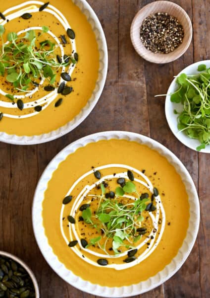 Two bowls containing Thai coconut pumpkin soup garnished with pumpkin seeds, micro greens and a drizzle of sour cream.