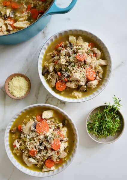 Two bowl of chicken wild rice soup topped with grated Parmesan cheese and thyme leaves. Surrounding the bowls are small bowls with cheese and thyme sprigs and a Dutch oven with the remainder of the soup in it.